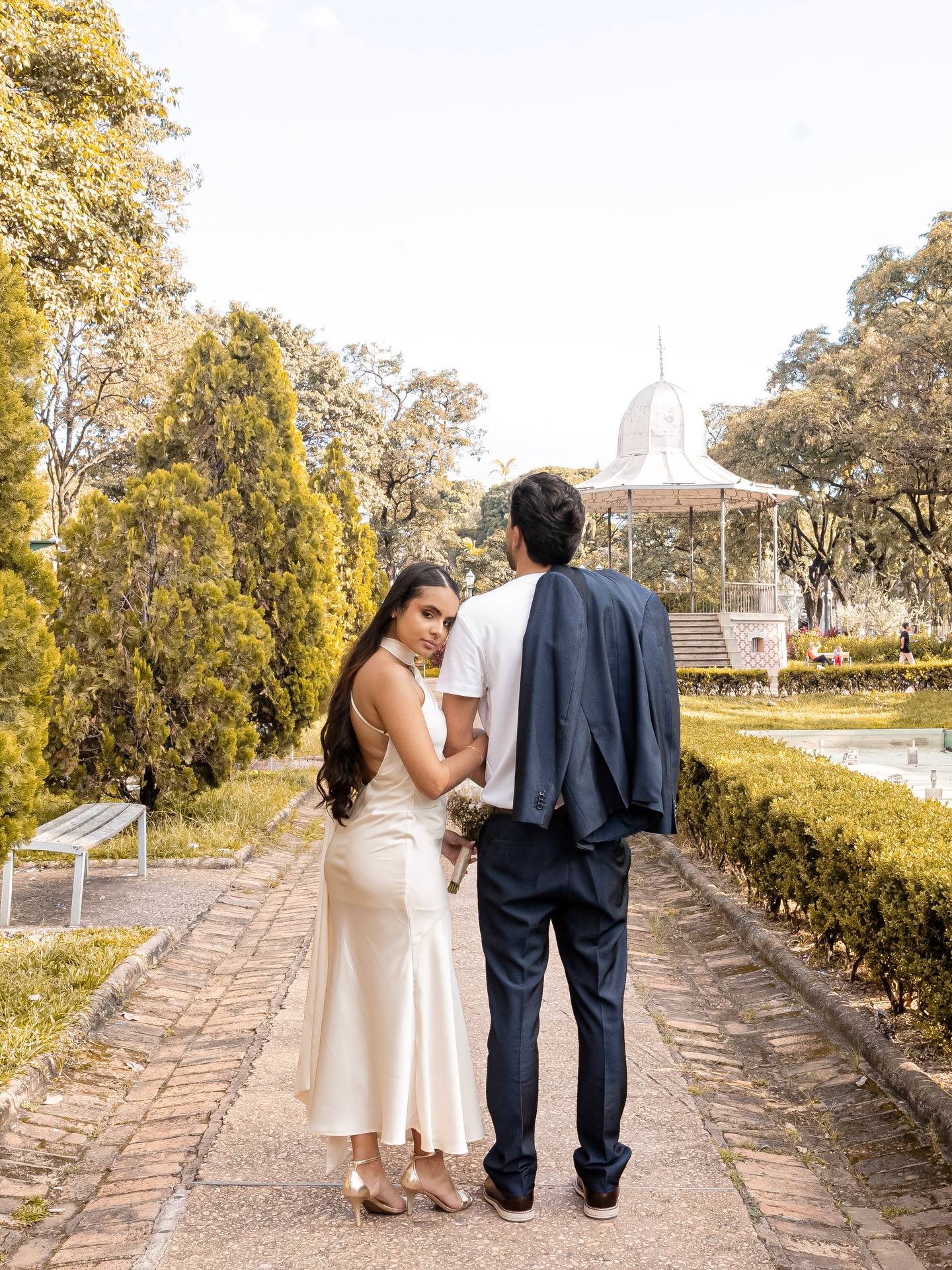 Casal caminhando de mãos dadas por praça arborizada com coreto