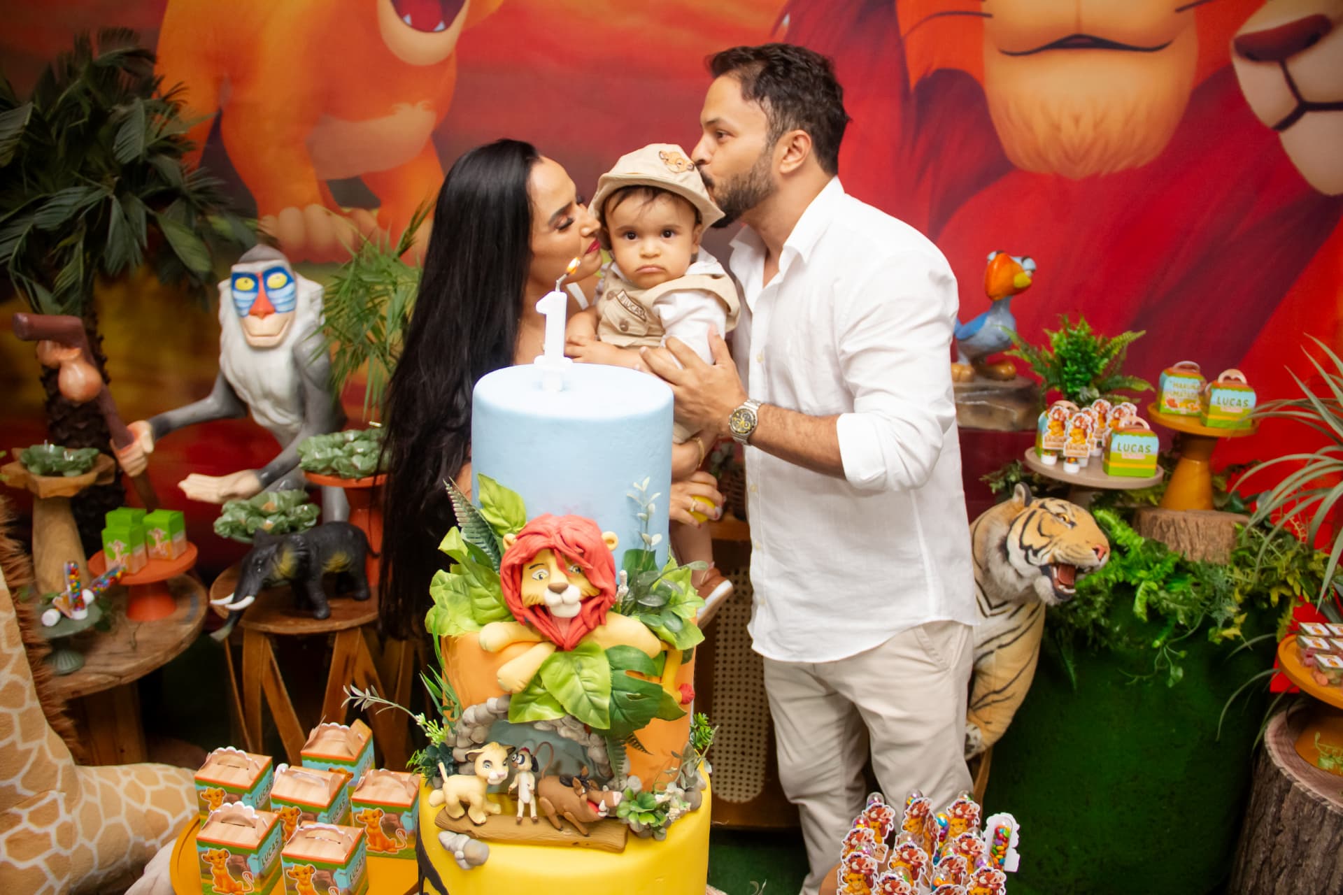 Pais beijando o bebê ao lado do bolo em festa de 1 ano com tema safári